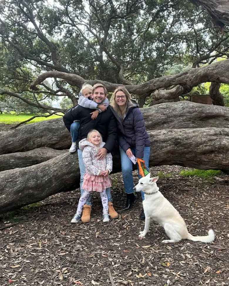 Isaac with his family and their white dog under a big old pōhutukawa tree.